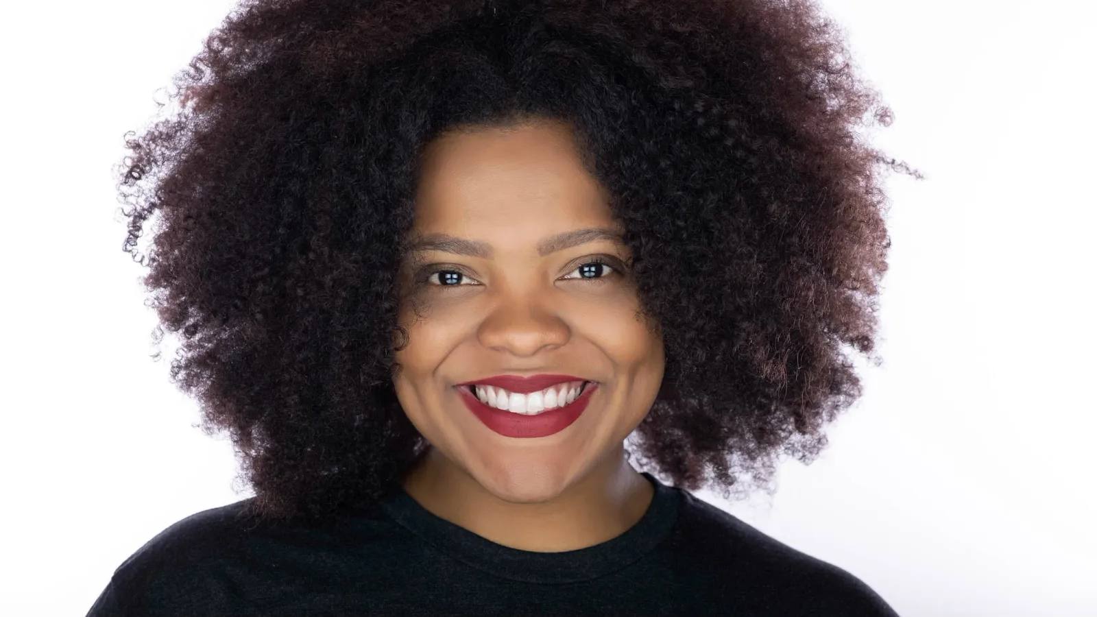 Headshot of a woman with full curly hair and red lipstick, smiling widely, in a black top, on a white background