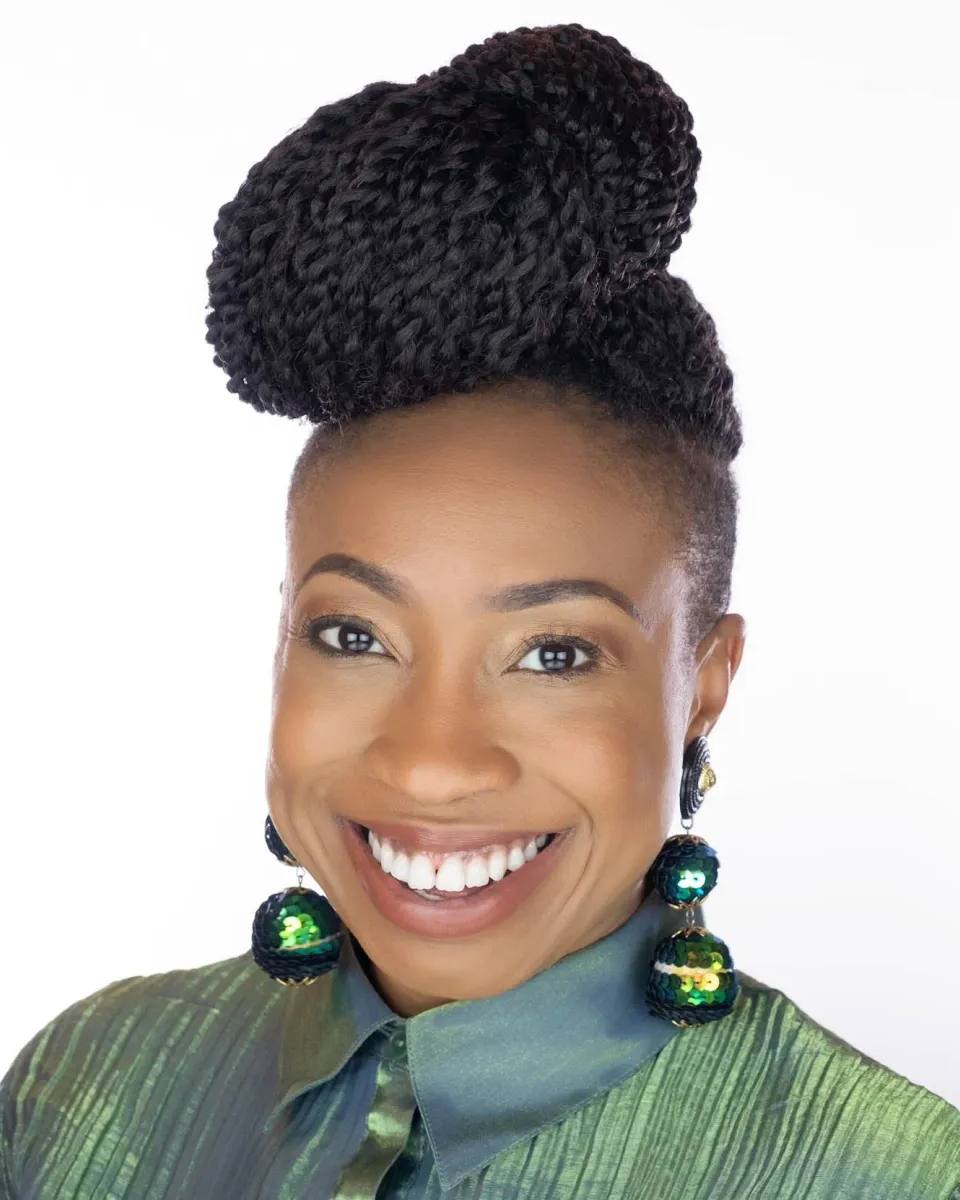 Headshot of a woman with braided hair in a high bun and large green sequin earrings, smiling widely, in a green collared shirt, on a white background