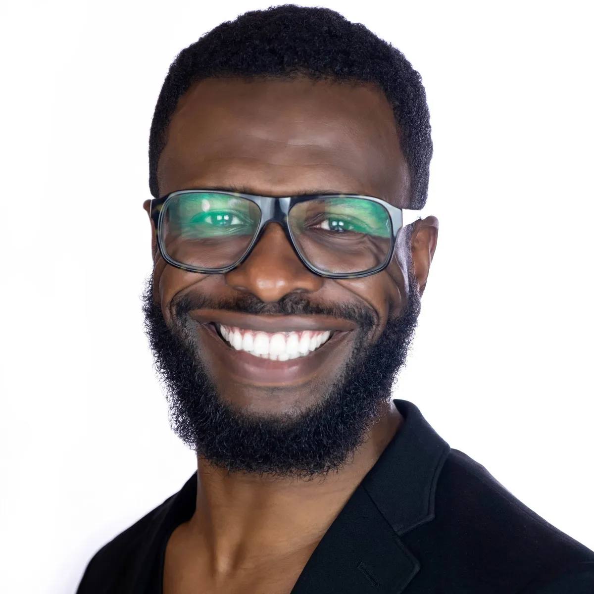 Headshot of a man with glasses and a full beard, smiling widely, in a black blazer, on a white background