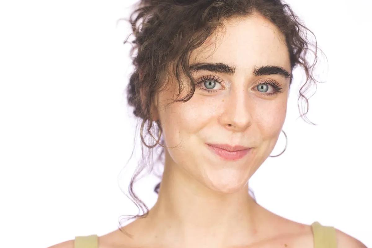 Headshot of a woman with curly dark hair pinned up and hoop earrings, slight smile, in an olive tank top, on a white background