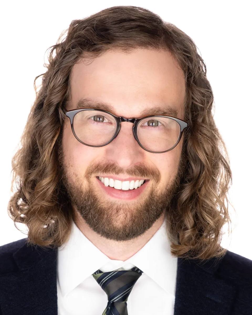 Headshot of a man with shoulder-length curly hair, glasses and a beard, smiling, in a navy suit, white shirt and striped tie, on a white background