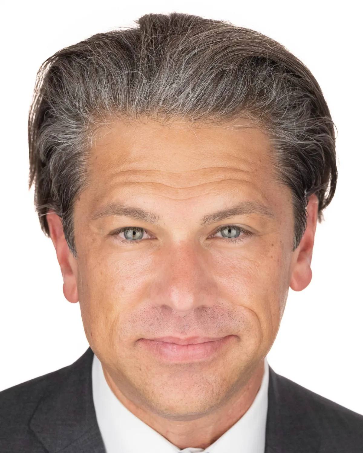 Close headshot of a man with swept-back greying hair, slight smile, in a charcoal suit, white shirt and light blue striped tie, on a white background
