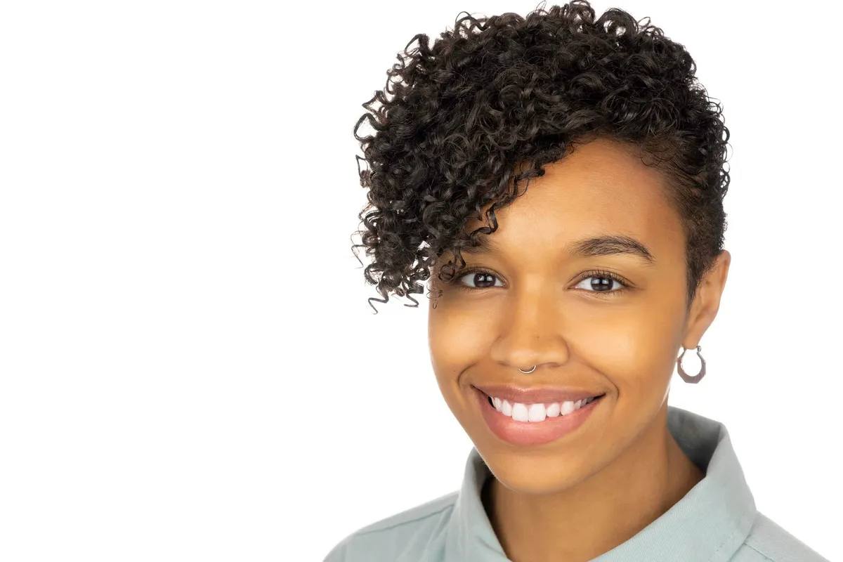 Headshot of a woman with short curly hair swept to one side, a septum ring and an earring, smiling, in a pale green collared shirt, on a white background