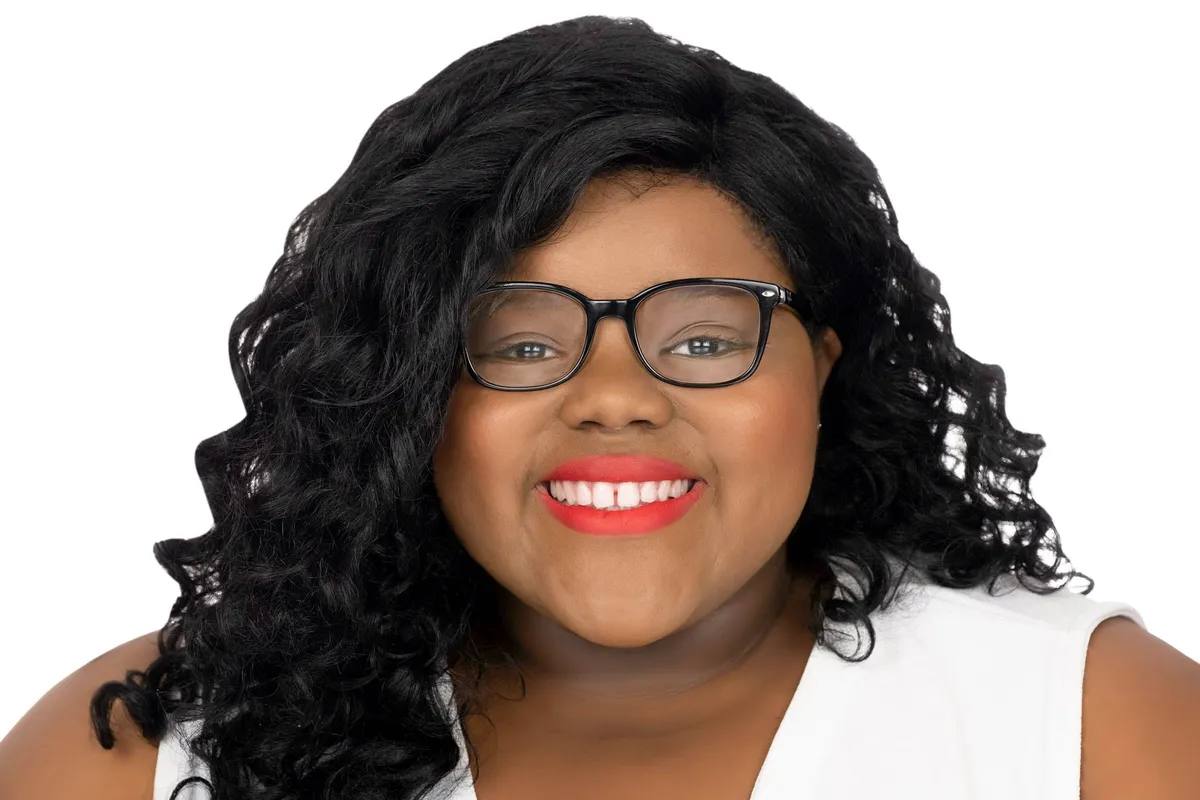 Headshot of a woman with long curly black hair, black glasses and red lipstick, smiling, in a white sleeveless top, on a white background