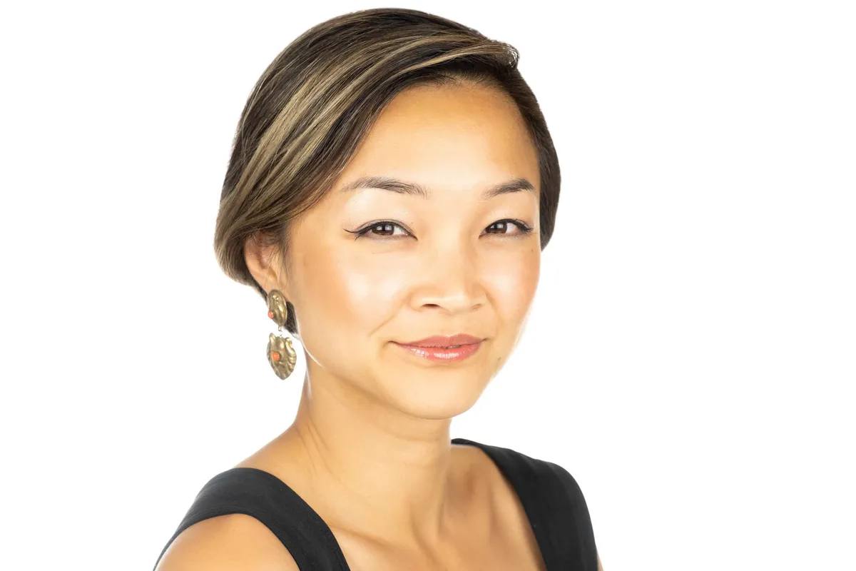 Headshot of a woman with short highlighted hair and a large drop earring, soft smile, in a black sleeveless top, on a white background