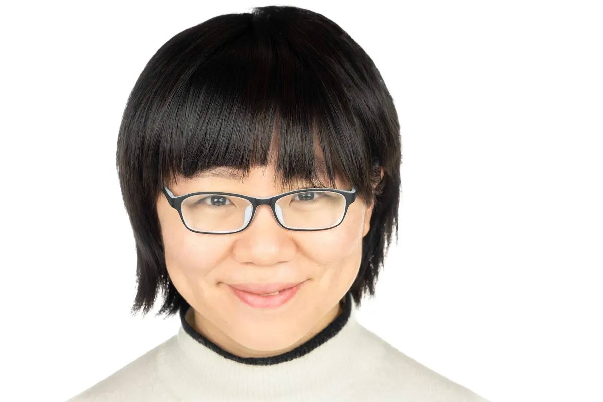 Headshot of a woman with a black bob and fringe and black glasses, smiling, in a cream turtleneck, on a white background