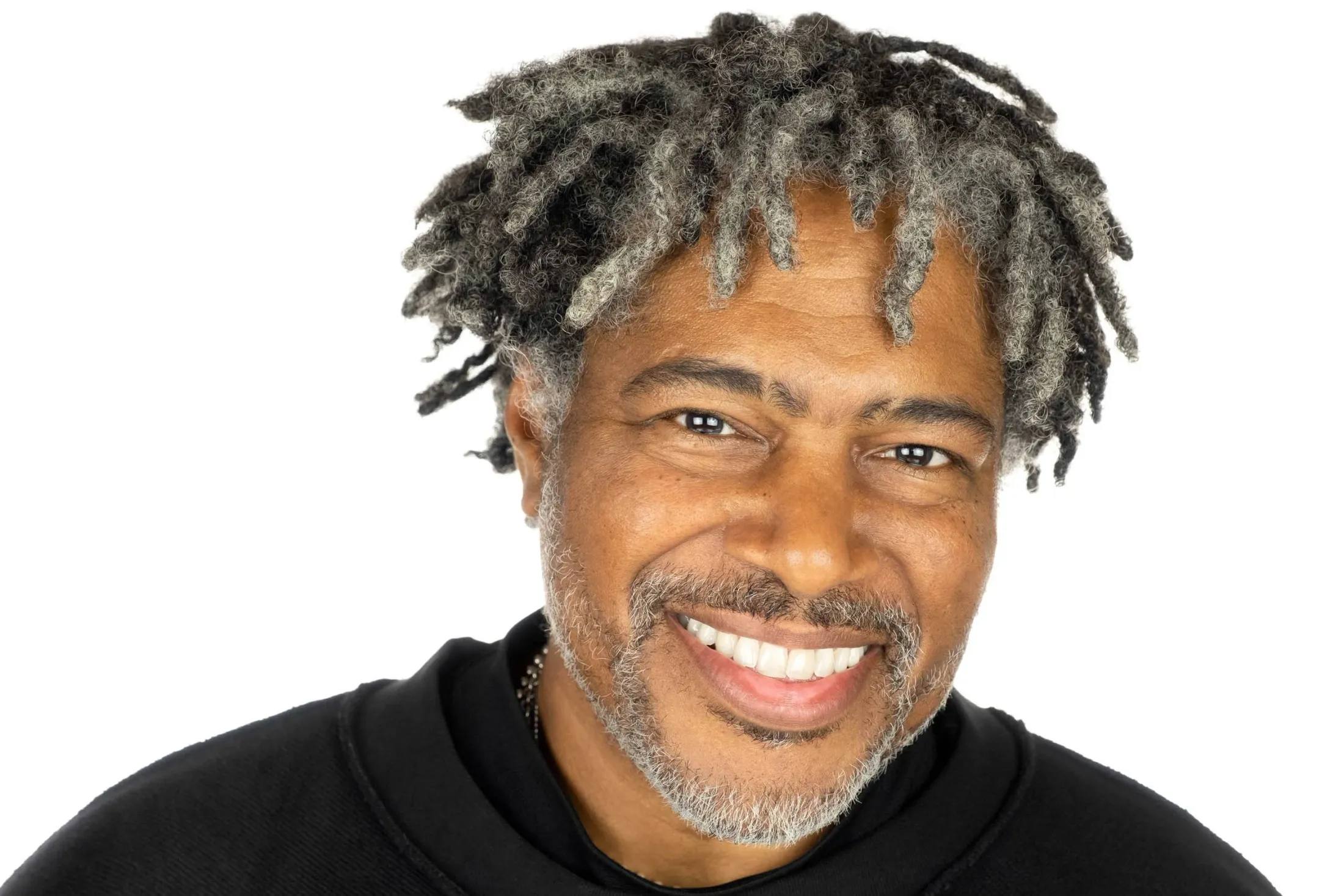 Headshot of a man with grey locs and a grey beard, smiling widely, in a black hooded top, on a white background
