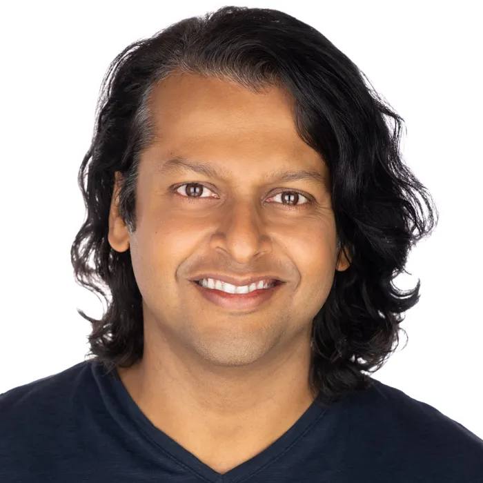 Headshot of a man with shoulder-length wavy black hair, smiling, in a navy V-neck top, on a white background