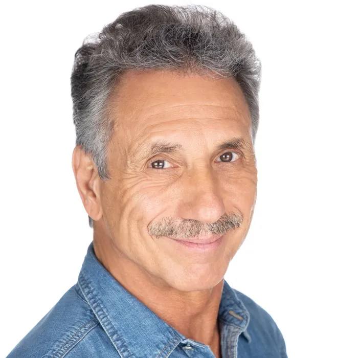Headshot of a man with grey hair and a grey moustache, head tilted, slight smile, in a denim shirt, on a white background